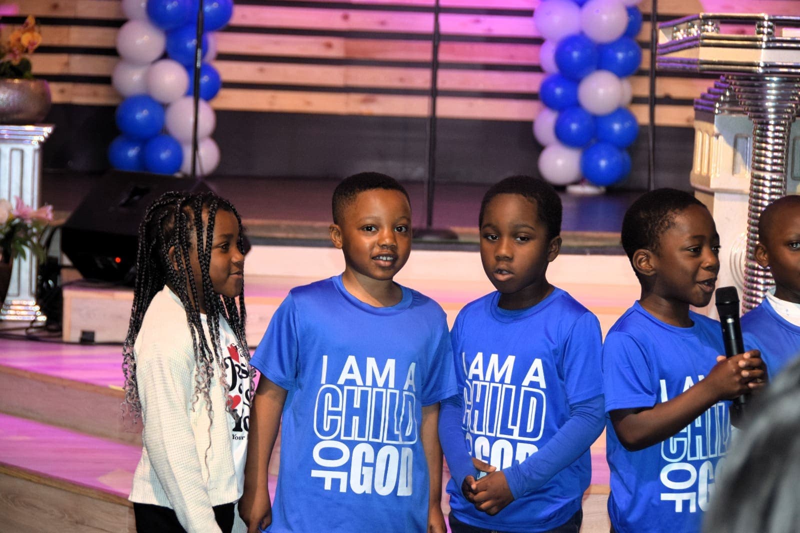 Four young children in blue holding microphones, blue balloons behind them